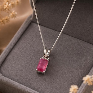 May include: A silver necklace with a rectangular, pink gemstone pendant. The pendant is set in a silver frame and hangs from a delicate silver chain. The necklace is displayed in a gray jewelry box.