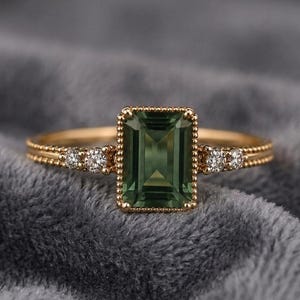 May include: A gold ring featuring a rectangular, emerald-cut green gemstone. The ring has small, clear gemstones on either side of the main stone and a beaded design along the band. The ring is a beautiful piece of jewellery.