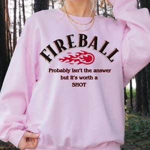 May include: Pink sweatshirt with the text "FIREBALL" in black with a red and orange flame graphic. The text "Probably Isn't the answer but it's worth a SHOT" is printed below the graphic.