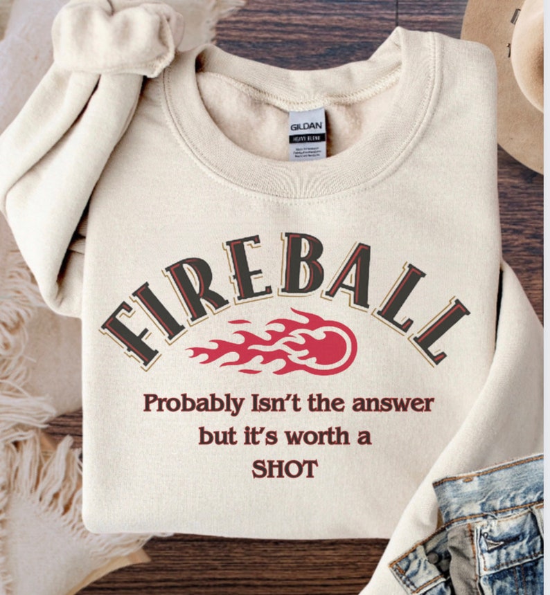 May include: A beige sweatshirt with the text "FIREBALL" in a brown and black font. The text is arched over a red and orange flame graphic. Below the graphic is the text "Probably Isn't the answer but it's worth a SHOT" in a red font.