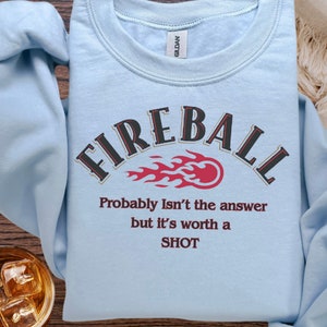 May include: Light blue sweatshirt with the text "FIREBALL" in a black and gold arch with a red flame graphic. The text "Probably Isn't the answer but it's worth a SHOT" is printed below the graphic.