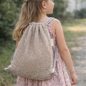 Könnte beinhalten: Ein beigefarbener Rucksack mit Kordelzug und Textur mit grauen Trägern. Der Rucksack ist aus einem weichen, plüschigen Material gefertigt. Das Bild zeigt eine junge Person, die den Rucksack mit einem Blumenkleid trägt.