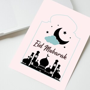 May include: A pink card with a white border and the text "Eid Mubarak" in brown lettering. The card features a black silhouette of a mosque with a crescent moon and stars in the sky.