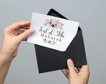 May include: A white rectangular greeting card featuring the black text Eid al-Fitr Mubarak centered between two watercolor floral arrangements in shades of pink, blue, and green, partially inserted into a solid black envelope.