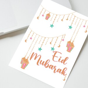 May include: A white card with a gold and pink string of lights with stars and crescent moons. The text "Eid Mubarak" is written in a pink and orange font.