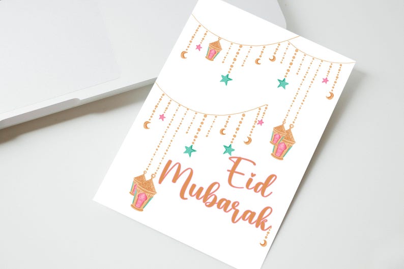May include: A white card with a gold and pink string of lights with stars and crescent moons. The text "Eid Mubarak" is written in a pink and orange font.