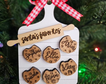 May include: A white wooden cutting board ornament with a red and white checkered ribbon. The board has a rolling pin with the text "santa's favorites" and six wooden cookie ornaments with names on them: Sophia, Ellie, Aria, Lincoln, Mpers, and Kitty.