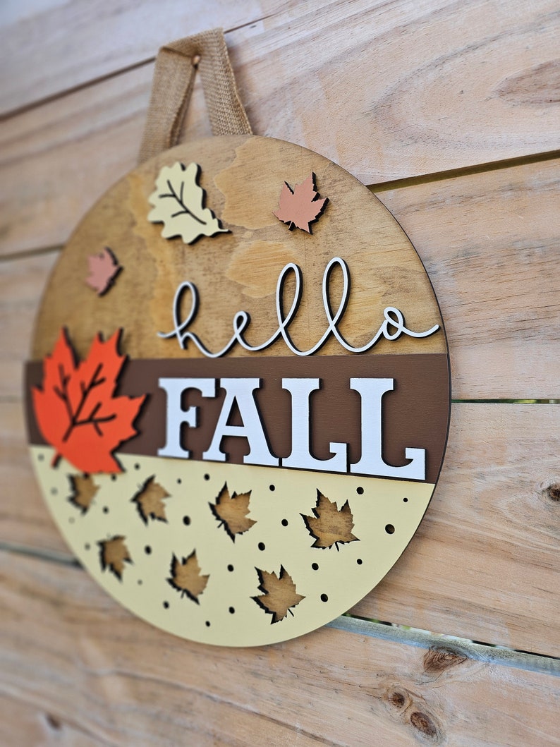 May include: A round wooden sign with the words 'hello fall' in brown script. The sign is decorated with fall leaves in orange, yellow, and brown. The sign is hanging on a wooden wall.