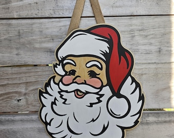 May include: Wooden Santa Claus face decoration with a red hat, white beard, and pink cheeks. The Santa face is outlined in black and has a burlap loop for hanging. A festive Christmas decoration.