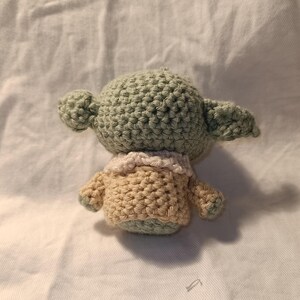 Pu&ograve; includere: Un giocattolo di peluche verde e beige all'uncinetto che ricorda un personaggio della saga di Star Wars. Il giocattolo ha una testa grande con una piccola bocca e grandi orecchie.