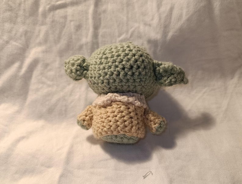 Pu&ograve; includere: Un giocattolo di peluche verde e beige all'uncinetto che ricorda un personaggio della saga di Star Wars. Il giocattolo ha una testa grande con una piccola bocca e grandi orecchie.