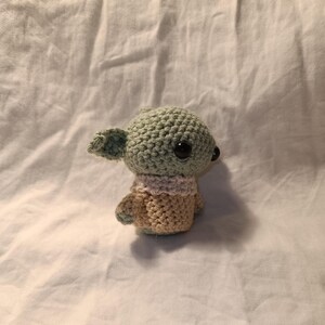Pu&ograve; includere: Un giocattolo di peluche verde e beige all'uncinetto con grandi occhi neri. Il giocattolo ha la forma di una piccola creatura con grandi orecchie e una piccola bocca.