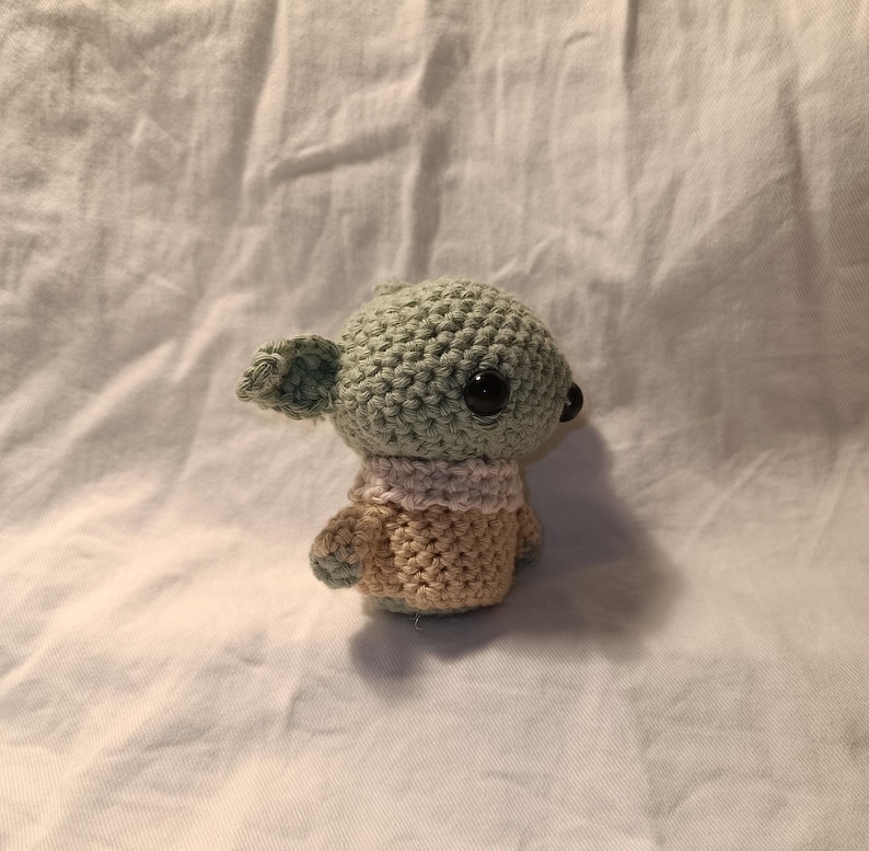 Pu&ograve; includere: Un giocattolo di peluche verde e beige all'uncinetto con grandi occhi neri. Il giocattolo ha la forma di una piccola creatura con grandi orecchie e una piccola bocca.
