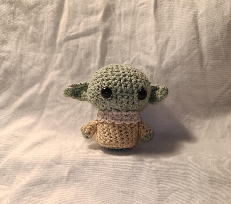 Pu&ograve; includere: Un giocattolo di peluche all'uncinetto di un personaggio alieno verde con occhi grandi e un maglione beige. Il giocattolo &egrave; seduto su uno sfondo di tessuto bianco.