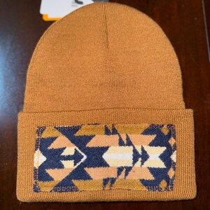 Puede incluir: Un gorro marr&oacute;n tejido con un parche rectangular con un dise&ntilde;o geom&eacute;trico en azul marino, beige y blanco. El gorro tiene un borde doblado.