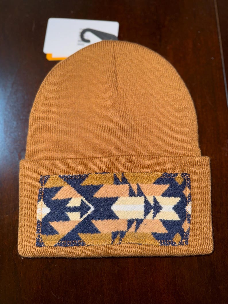 Puede incluir: Un gorro marr&oacute;n tejido con un parche rectangular con un dise&ntilde;o geom&eacute;trico en azul marino, beige y blanco. El gorro tiene un borde doblado.