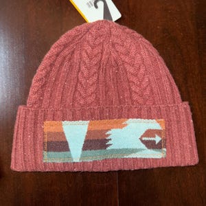 Puede incluir: Un gorro de punto rojo ladrillo con un dise&ntilde;o de punto trenzado. El gorro presenta un parche rectangular con un dise&ntilde;o geom&eacute;trico en tonos naranja, marr&oacute;n y verde azulado. Una etiqueta blanca con un gancho negro est&aacute; adherida al gorro.