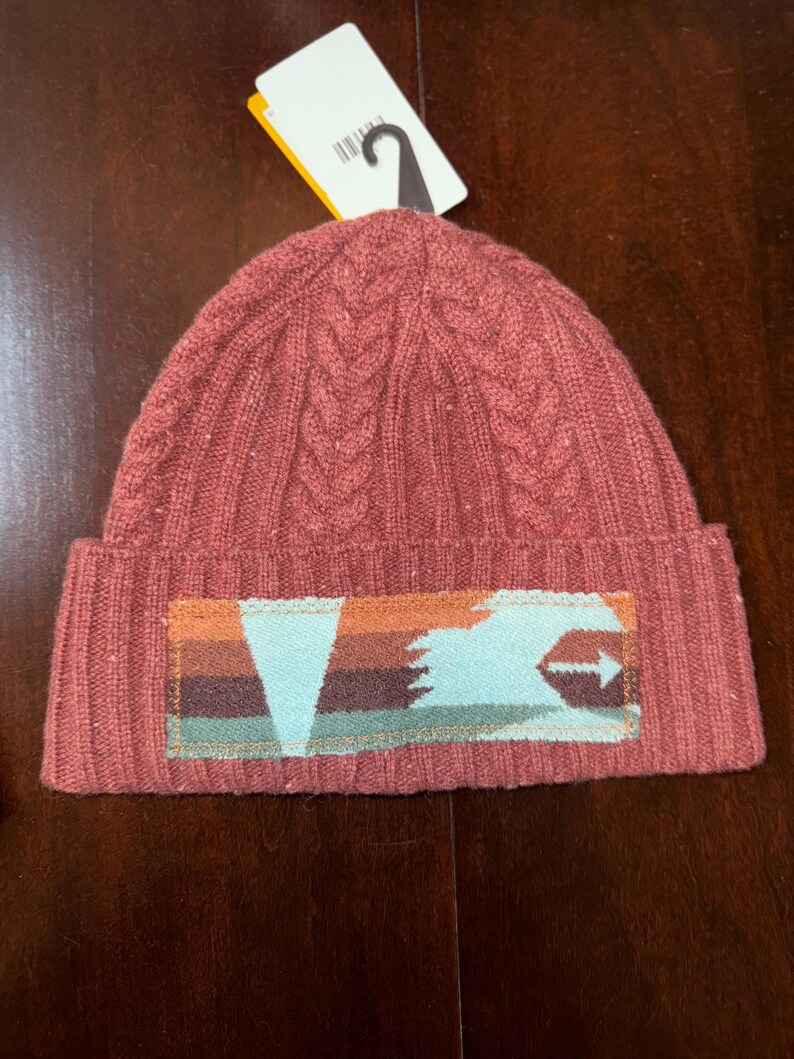 Puede incluir: Un gorro de punto rojo ladrillo con un dise&ntilde;o de punto trenzado. El gorro presenta un parche rectangular con un dise&ntilde;o geom&eacute;trico en tonos naranja, marr&oacute;n y verde azulado. Una etiqueta blanca con un gancho negro est&aacute; adherida al gorro.
