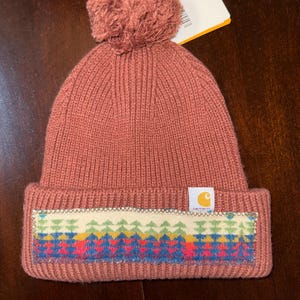 Puede incluir: Un gorro de punto rosa polvoriento con un pomp&oacute;n a juego. El gorro presenta un dise&ntilde;o geom&eacute;trico colorido en el borde y una etiqueta con el logotipo de Carhartt. El gorro est&aacute; hecho de punto acanalado.