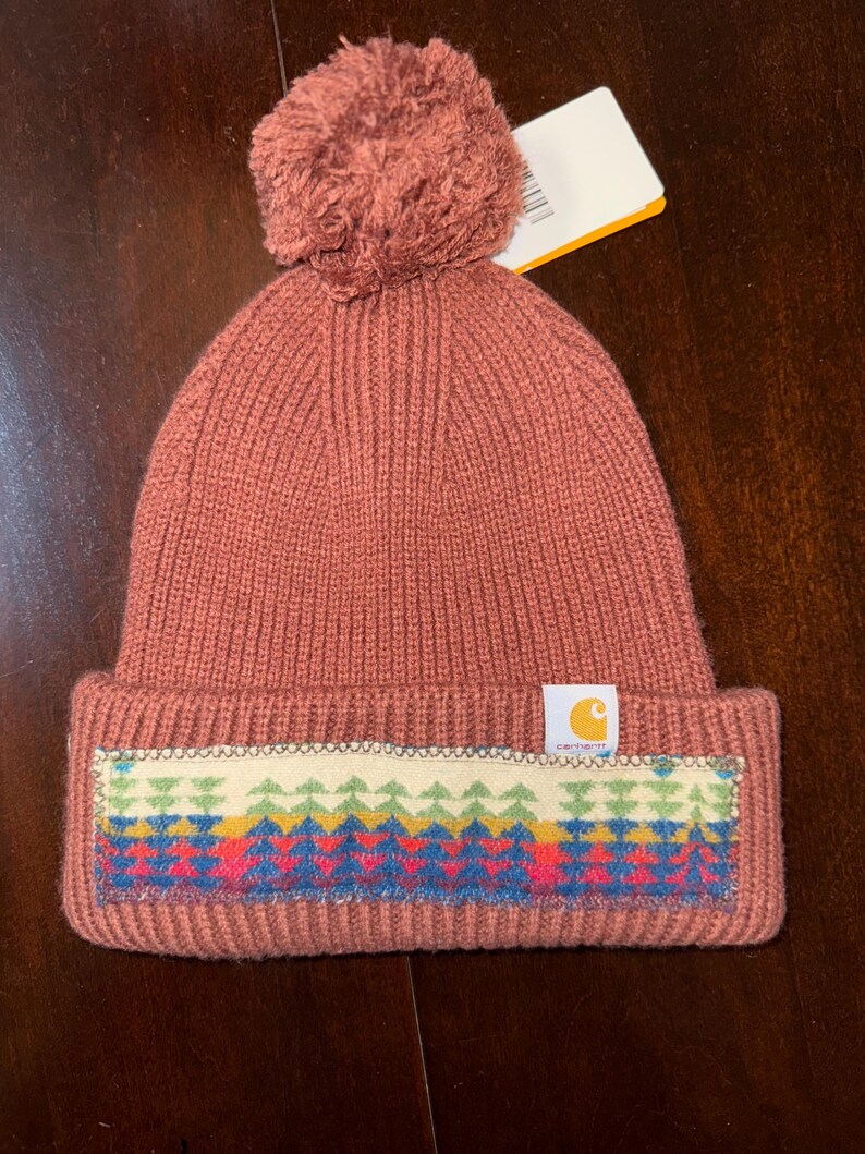 Puede incluir: Un gorro de punto rosa polvoriento con un pomp&oacute;n a juego. El gorro presenta un dise&ntilde;o geom&eacute;trico colorido en el borde y una etiqueta con el logotipo de Carhartt. El gorro est&aacute; hecho de punto acanalado.