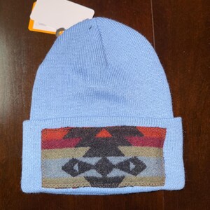 Puede incluir: Un gorro azul claro con un borde doblado. El borde presenta un dise&ntilde;o geom&eacute;trico en tonos rojos, naranjas, marrones, grises y negros. El gorro est&aacute; hecho de un material suave y tejido.