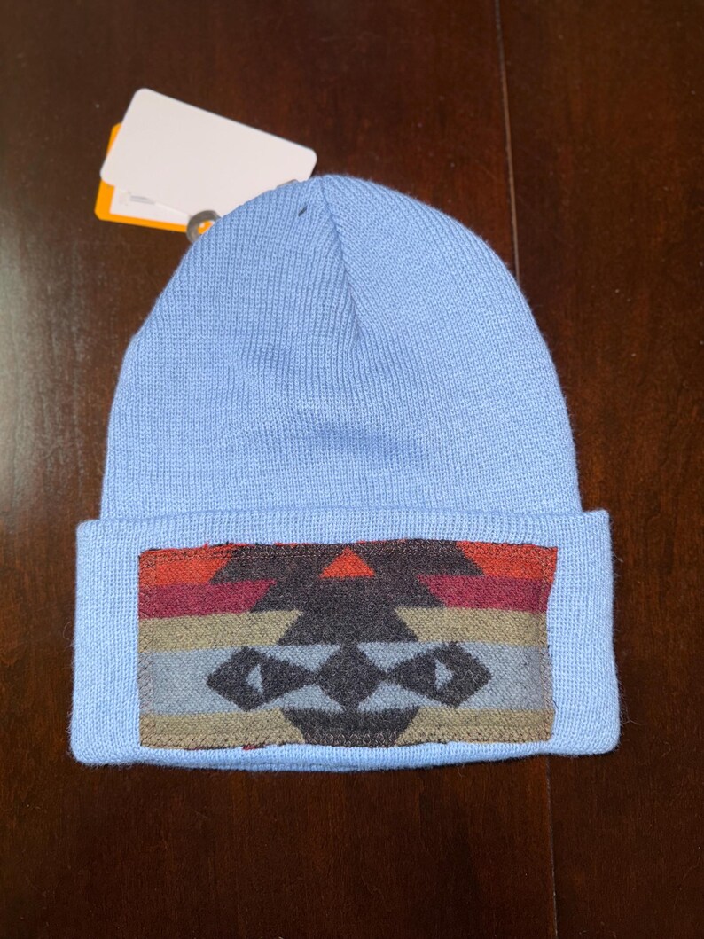 Puede incluir: Un gorro azul claro con un borde doblado. El borde presenta un dise&ntilde;o geom&eacute;trico en tonos rojos, naranjas, marrones, grises y negros. El gorro est&aacute; hecho de un material suave y tejido.