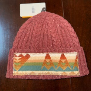 Puede incluir: Un gorro de punto en un tono marr&oacute;n rojizo apagado. El gorro presenta un dise&ntilde;o de punto trenzado y un parche rectangular con un patr&oacute;n geom&eacute;trico colorido en tonos naranja, verde y crema. Una etiqueta est&aacute; adjunta.