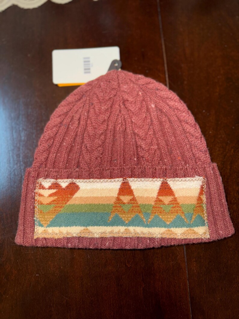 Puede incluir: Un gorro de punto en un tono marr&oacute;n rojizo apagado. El gorro presenta un dise&ntilde;o de punto trenzado y un parche rectangular con un patr&oacute;n geom&eacute;trico colorido en tonos naranja, verde y crema. Una etiqueta est&aacute; adjunta.