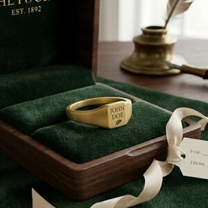 Könnte beinhalten: Ein goldener Siegelring mit der Gravur "JOHN DOE" und einem kleinen Blattdesign. Der Ring liegt in einer dunkelgrünen, samtgefütterten Schatulle. Die Schatulle ist geöffnet und zeigt den Ring und das Samtinterieur.