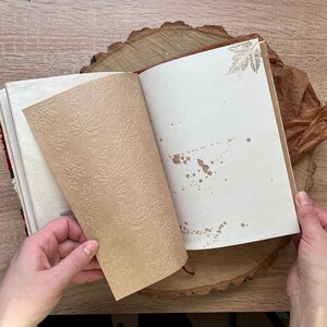 May include: An open journal with brown paper pages featuring a leaf pattern. The pages are blank and have brown splatter paint. The journal is on a wooden surface.