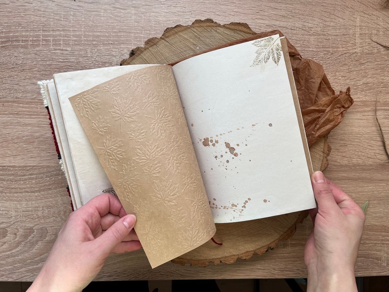 May include: An open journal with brown paper pages featuring a leaf pattern. The pages are blank and have brown splatter paint. The journal is on a wooden surface.