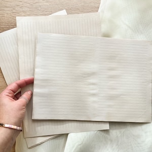 May include: A stack of lined paper sheets, with one sheet folded in half to create a notebook. The paper is a light beige color and the lines are a darker beige color.