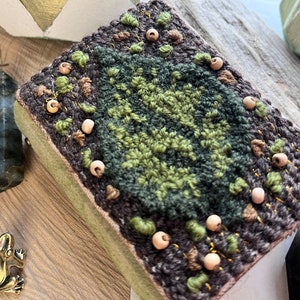 May include: A small, square, green and brown book with a woven design and wooden beads around the edges. The book is sitting on a wooden surface.