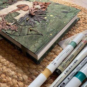 May include: A handmade journal with a green fabric cover, adorned with embroidered leaves, a mushroom design, and gold beads. Several art markers and a paintbrush are arranged beside the journal on a woven mat, suggesting a craft or art theme.