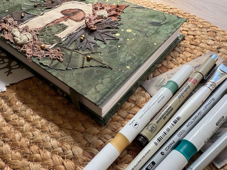 May include: A handmade journal with a green fabric cover, adorned with embroidered leaves, a mushroom design, and gold beads. Several art markers and a paintbrush are arranged beside the journal on a woven mat, suggesting a craft or art theme.