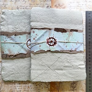 May include: Two sheets of light green textured paper with brown edges, tied together with twine. The paper has a distressed look with brown paint splatters and a decorative sticker of a ship's wheel with flowers.