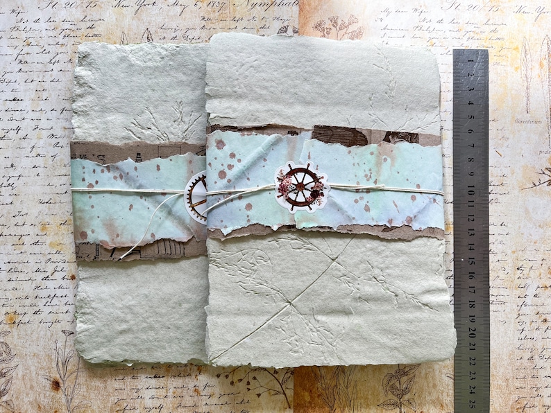 May include: Two sheets of light green textured paper with brown edges, tied together with twine. The paper has a distressed look with brown paint splatters and a decorative sticker of a ship's wheel with flowers.