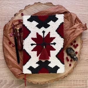 May include: A white and red needlepoint journal cover with a black geometric pattern and a red tassel. The cover is on a wooden surface with a brown paper background.