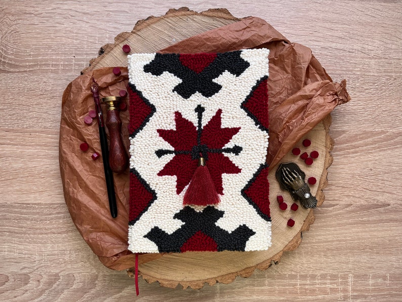 May include: A white and red needlepoint journal cover with a black geometric pattern and a red tassel. The cover is on a wooden surface with a brown paper background.