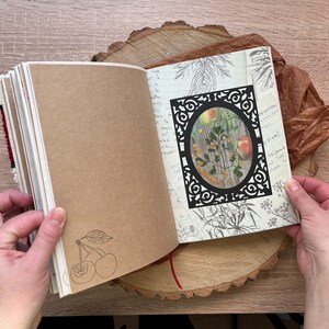 May include: Open journal with brown paper pages. The left page has a hand-drawn sketch of two cherries with a stem and leaf. The right page has a black decorative frame with a vintage botanical illustration of a plant with yellow flowers and red apples inside. The journal is on a wooden surface.