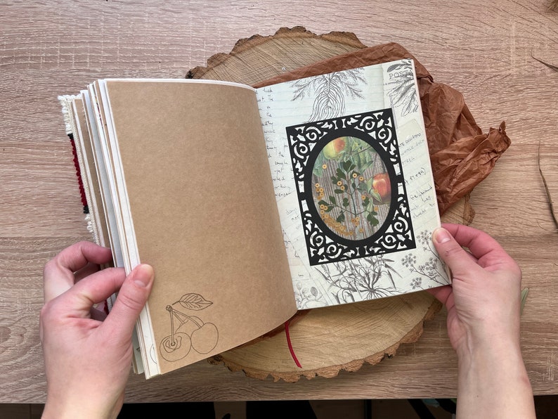 May include: Open journal with brown paper pages. The left page has a hand-drawn sketch of two cherries with a stem and leaf. The right page has a black decorative frame with a vintage botanical illustration of a plant with yellow flowers and red apples inside. The journal is on a wooden surface.
