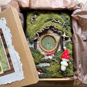 May include: A handmade journal with a green and brown felt cover depicting a hobbit hole with a round door. The journal comes with a crocheted mushroom bookmark with a red cap and white stem.