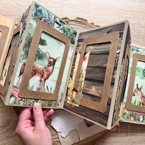 May include: A brown and beige scrapbook with multiple photo frames. The frames are decorated with stitching and have a rustic woodgrain background. The scrapbook features images of a deer in a forest setting.