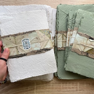 May include: Two stacks of handmade paper, one white and one green, with decorative paper and twine accents. The paper is textured and has a rustic look.