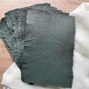 May include: A pile of handmade, dark grey paper sheets with rough, textured surfaces. The paper has deckled edges and is displayed on a light wooden surface with white fabric in the background. Suitable for art projects.