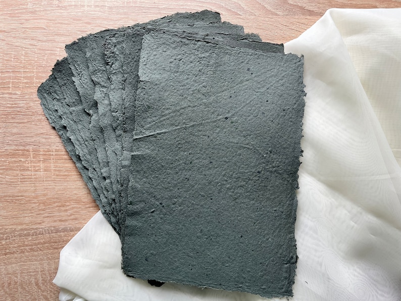 May include: A pile of handmade, dark grey paper sheets with rough, textured surfaces. The paper has deckled edges and is displayed on a light wooden surface with white fabric in the background. Suitable for art projects.