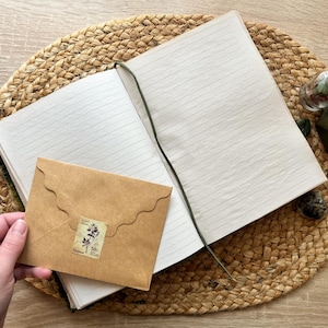 K&ouml;nnte beinhalten: Ein braunes, ledergebundenes Tagebuch mit linierten Seiten, ein brauner Umschlag mit dekorativem Rand und einer Vintage-Briefmarke sowie eine kleine Glasflasche mit getrockneten Blumen darin. Das Tagebuch liegt auf einer geflochtenen Strohmatte.