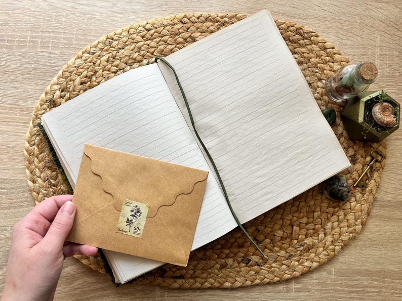 K&ouml;nnte beinhalten: Ein braunes, ledergebundenes Tagebuch mit linierten Seiten, ein brauner Umschlag mit dekorativem Rand und einer Vintage-Briefmarke sowie eine kleine Glasflasche mit getrockneten Blumen darin. Das Tagebuch liegt auf einer geflochtenen Strohmatte.