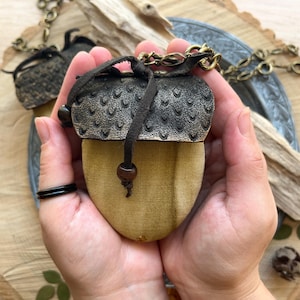 Op de afbeelding: Een klein, eikelvormig tasje met een bruin leren klepje en een ketting. Het tasje is gemaakt van lichtbruine stof en heeft een kralenkwast.