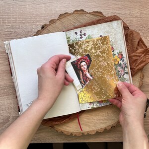 May include: A person is holding a gold leaf sheet over a page in a journal. The page has a floral design and a picture of a woman wearing a red and gold crown.